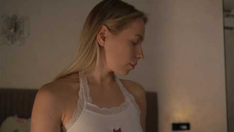 Video of a fair-skinned, blonde woman in a white lace-trimmed camisole, gazing thoughtfully in a dimly lit bedroom with neutral walls and a nightstand lamp.