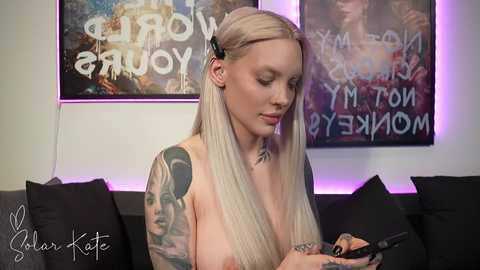 Video of a topless, tattooed, platinum blonde woman holding a cell phone. She has a slender physique, light skin, and piercings. Background features abstract art and purple lighting.