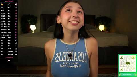 Video of a smiling teenage girl with light skin and straight brown hair, wearing a blue tank top with white text. Background includes a dimly lit bedroom with a bed and potted plants.