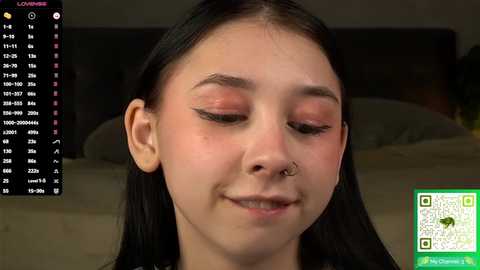 Video of a young Asian woman with straight black hair, wearing a nose ring and light makeup, smiling softly. Background shows a bed and a smartphone displaying a \"LOV\" app.