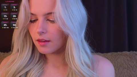 Video of a young, fair-skinned woman with long platinum blonde hair, wearing a nose ring and light makeup, sitting on a beige couch.