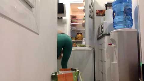 A video of a person wearing teal leggings and a green shirt, bending over to retrieve an orange bag from a white door, in a narrow kitchen with white cabinets, a refrigerator, and a water dispenser.