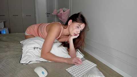 Video of a young woman with long brown hair, lying on a bed, typing on a white keyboard, wearing a pink tank top and shorts, in a modern bedroom.