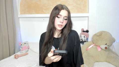 Video of a young woman with long, straight brown hair, wearing a black T-shirt, holding a black phone, surrounded by plush toys in a cozy, pastel-toned bedroom.