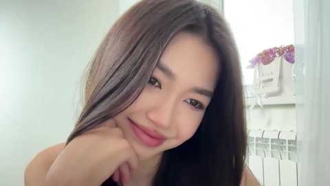 Video of an Asian woman with long, straight black hair, light skin, and a slim figure, smiling softly, wearing a black top, standing in a bright, white-walled room with a radiator and a floral arrangement in the background.