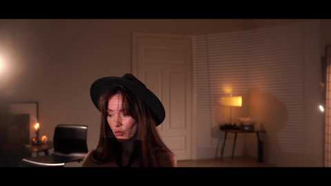 A video captures a woman with long brown hair, wearing a black witch hat, indoors. She's in a dimly lit room with white blinds, a lamp, and a desk.