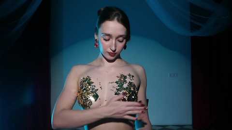 Video of a topless woman with fair skin and brown hair, adorned with glittering gold and green body paint on her chest, standing in a dimly lit, draped room, wearing red earrings.