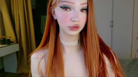 Video of a pale-skinned woman with long, straight, fiery red hair and bold, dramatic makeup, including winged eyeliner and red lipstick, wearing a white choker necklace. Background shows a beige curtain and a white wardrobe.