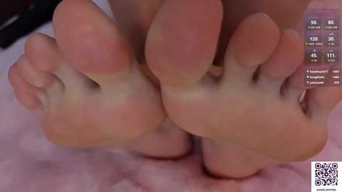 Video of two clean, light-skinned feet, toes slightly curled, resting on a pink, plush surface. Background includes a blurred, dark-colored object and a QR code in the bottom right corner.