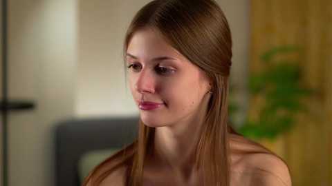 Video of a fair-skinned, slender young woman with straight, light brown hair, wearing a red lip color, in a casual indoor setting with wooden furniture and green plants in the background.