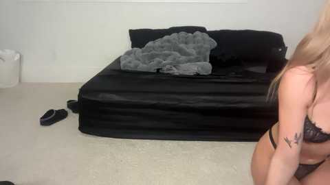 Video of a woman with light skin and blonde hair, wearing black lace lingerie, standing beside a bed with black sheets and a gray blanket, in a minimalist, white-walled room.