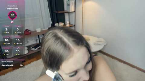 Video of a young woman with light brown hair, wearing glasses, lying on a white carpet in a modern bedroom. The room has a white wall, wooden floor, and a shelf with books and a lamp.