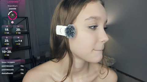 Video of a young Caucasian woman with light skin, brown hair, and a white, fluffy makeup sponge on her forehead, smiling. Background includes a black metal shelf with pink and white objects.
