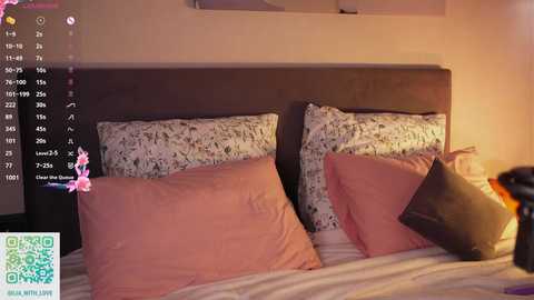 Video of a cozy bedroom with a beige headboard, two pink pillows, and a floral-patterned pillow on a white bed. A green and yellow pillow is partially visible. The room has a warm, inviting atmosphere.