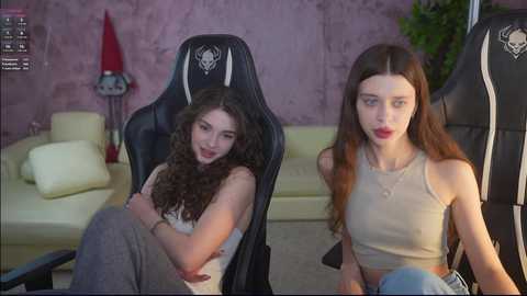 Video of two young women in a cozy living room. One is curly-haired, wearing a tank top, sitting on a gaming chair. The other has long straight hair, wearing a crop top, seated on a couch.