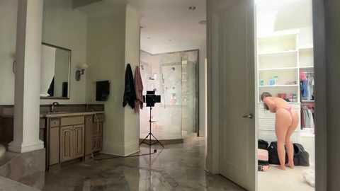 Video of a modern, spacious bathroom with a nude woman bending over, wearing a pink thong, facing a mirror. The room features a sleek, white vanity and a glass-enclosed shower.
