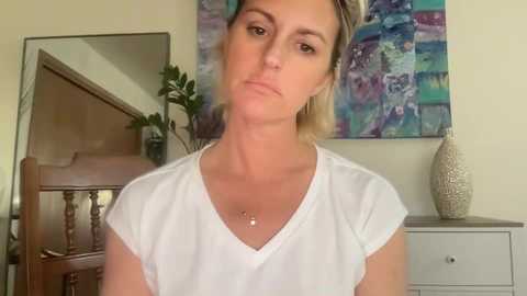 Video of a blonde woman with a neutral expression, wearing a white V-neck T-shirt, standing in front of a colorful abstract painting and a white dresser with a vase.