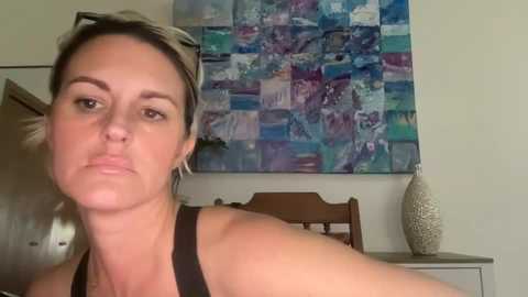 A video of a woman with short blonde hair, wearing sunglasses and a black tank top, standing in front of a colorful abstract painting and a beige vase.