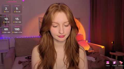 Video of a young Caucasian woman with long, wavy red hair, fair skin, and closed eyes, sitting on a bed in a dimly lit room with a glowing candle on a side table.