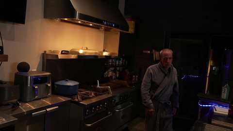 Video of a dimly lit, rustic kitchen with a middle-aged man in a gray apron, standing next to a stove.