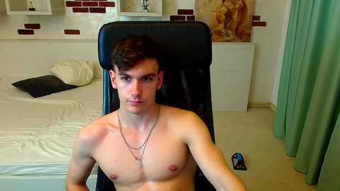 Video of a young, shirtless, pale-skinned man with short dark hair, sitting in a black leather chair in a modern bedroom. Background includes a bed, a green curtain, and a framed painting.