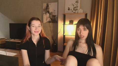 Video of two young women with fair skin and long black hair, one in a black top, sitting on a wooden table in a dimly lit room with beige walls and wooden shelves.