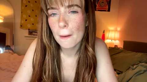 Video of a young woman with fair skin, long brown hair, and freckles, lying on a bed in a cozy, dimly-lit room with warm lighting, yellow wall art, and a green bedspread.