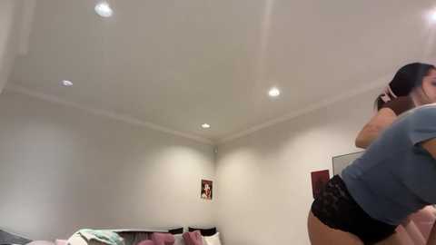 Video of a dimly lit bedroom with white walls, recessed ceiling lights, a bed with pink and white sheets, and a black-clad woman in the foreground, partially visible.