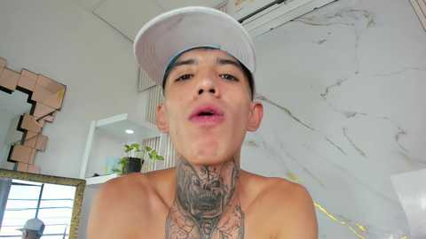Video of a shirtless young man with a white baseball cap, tattoos on his neck and chest, indoors with white marble walls and a modern, geometric mirror.