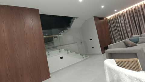 Modern, minimalist living room with a white sofa, wooden paneling, glass staircase, and beige carpet, lit by recessed ceiling lights and floor lamps.