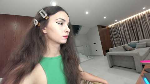 Video of a young woman with long brown hair, wearing a green sleeveless top, holding a hair dryer, standing in a modern living room with a gray sofa and brown curtains.