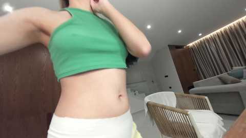 Video of a fair-skinned woman with a medium build, wearing a green crop top and white shorts, standing in a modern, minimalist living room with beige and wooden decor.