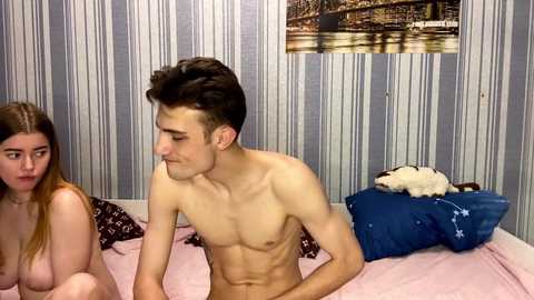 Video of a nude man with a lean build and short dark hair, sitting on a pink bed with a woman partially visible, both in a bedroom with striped wallpaper and a Paris-themed wall hanging.