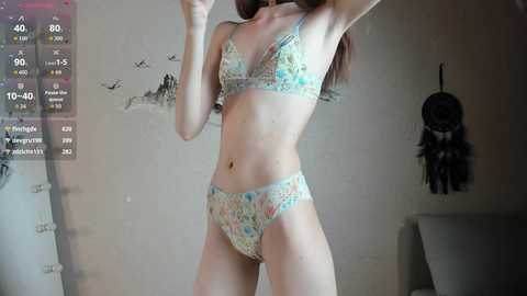 A video of a slender, fair-skinned woman in floral lingerie, standing in a dimly lit room with a dreamcatcher on the wall and a calendar displaying fitness stats.