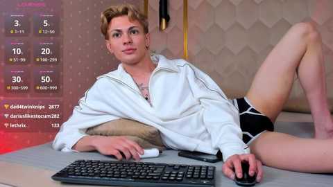 Video of a light-skinned, short-haired person in a white jacket, black shorts, lying on a bed with a computer keyboard, surrounded by social media stats and a geometric wallpaper.