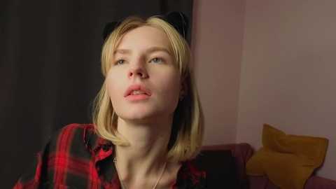 Video of a young, fair-skinned woman with straight blonde hair, wearing a red and black plaid shirt, and black cat ears, looking introspective against a pink and black background.