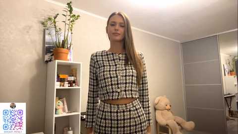 Video of a young woman with long, straight blonde hair wearing a black-and-white houndstooth checkset, standing in a cozy, minimalist room with beige walls, a potted plant, a bookshelf, and a teddy bear.