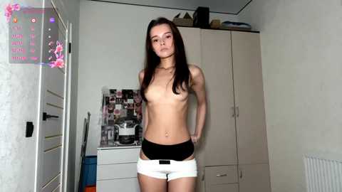 A video of a topless, slender Asian woman with long brown hair, wearing white panties, standing in a cluttered bedroom with white furniture and a bulletin board.