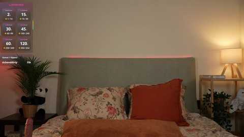 Video of a cozy bedroom with a teal headboard, floral-patterned bedding, orange throw pillows, a digital clock, a potted plant, and a table lamp.