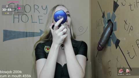 A video shows a blonde woman holding a blue sex toy over her mouth in a dimly lit room with a \"Glow Hole\" sign and a stuffed dildo on the wall.