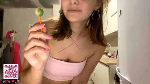 Video of a young woman with straight brown hair, wearing a pink crop top and large hoop earrings, holding a lollipop in a white kitchen.