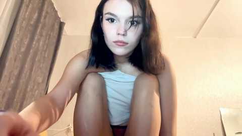 A video of a young, fair-skinned woman with shoulder-length black hair, wearing a white tank top, sitting in a dimly lit room with beige walls and a patterned curtain.