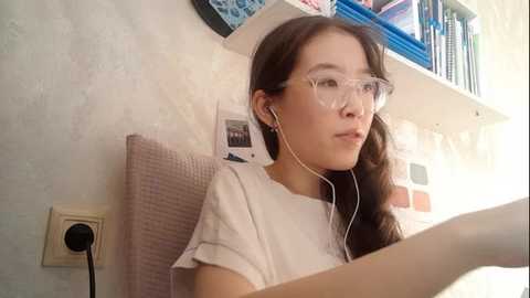 Video of an Asian woman with long brown hair, wearing clear glasses, white T-shirt, and white earbuds, sitting on a beige cushioned chair, surrounded by books and a wall-mounted shelf with a small framed photo.