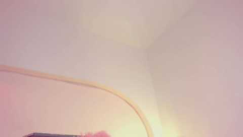 Video of a dimly lit room corner with a pale pink wall and a faintly visible, blurred object, possibly a piece of furniture, in the lower left. The room's ambiance is soft and muted, with a warm, diffused light.