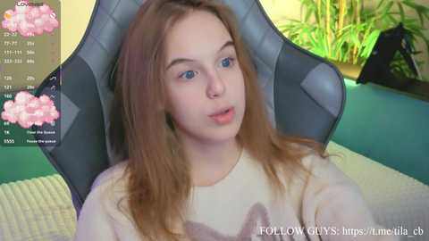 Video of a young woman with light skin, long brown hair, and blue eyes, wearing a white sweater, seated in a gray gaming chair, indoors with green plants and a blue background.