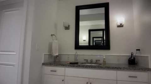 Video of a modern bathroom with a white granite countertop, black-framed mirror, wall-mounted sconces, and a white towel rack. The room is bright and clean, with a sleek, contemporary design.