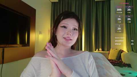 Video of an Asian woman with long brown hair, light skin, wearing a white off-shoulder top, smiling. She's in a modern living room with a TV and green curtains in the background.