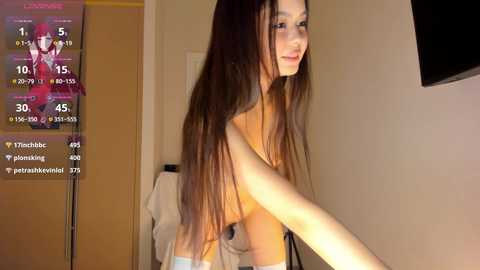 Video of a nude woman with long brown hair, slender physique, and small breasts, standing in a dimly lit room.