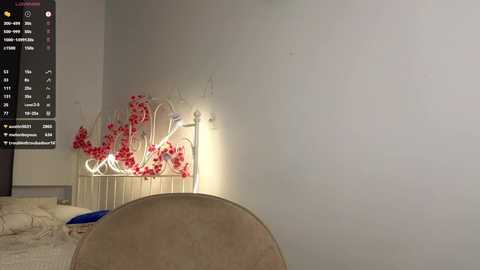 Video of a cozy bedroom corner with a beige cushioned headboard, a small table holding vibrant red flower decor, and a flat-screen TV displaying a digital clock and weather information.