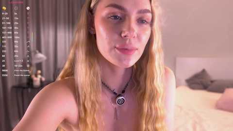 Video of a young blonde woman with wavy hair, wearing a black necklace, standing in a dimly lit bedroom.